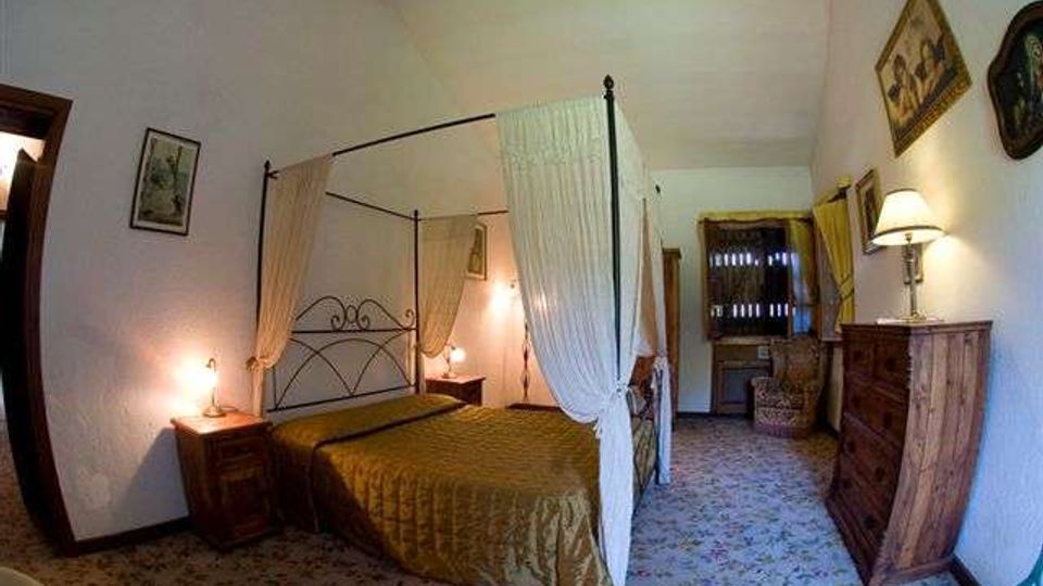 Camera da letto in stile rustico con letto a baldacchino in ferro e tende bianche, copriletto dorato e comodini con lampade ai lati. Pavimento piastrellato decorato, pareti bianche con quadri e mobili in legno, con una finestra in fondo alla stanza.