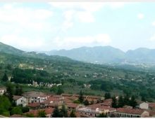 Veduta panoramica di un paese con case dai tetti in terracotta in primo piano, circondato da campi e boschi. Sullo sfondo si estendono colline e montagne sotto un cielo parzialmente nuvoloso.