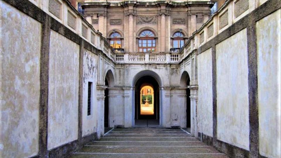 Passaggio pedonale in pietra tra alte pareti intonacate, che conduce a un portico con arcate. In fondo si vede la facciata di un edificio storico con finestre ad arco, colonne e balconata sopra un grande arco centrale.