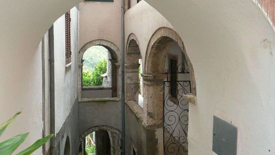 Passaggio stretto in un borgo, con scala in pietra e pavimento con muschio, coperto da un grande arco e affiancato da portici ad arcate. Sulle pareti intonacate si vedono una piccola balconata con ringhiera in ferro battuto e aperture ad arco che conducono a cortili interni.