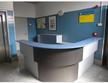 Reception di un ufficio con bancone curvo bianco e blu e pareti azzurre. Sullo sfondo si vedono una porta, un armadietto, quadri e una bacheca con orologio e dispositivi di sicurezza, con pavimento in graniglia.