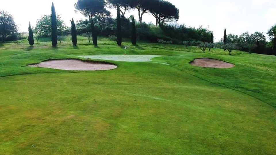 Green di un campo da golf su terreno ondulato, con due bunker di sabbia ai lati e una bandiera al centro. Sullo sfondo si vedono alberi alti e siepi che delimitano l’area.