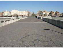 Ampio ponte pedonale lastricato in pietra, con parapetti in cemento e lampioni lungo i lati, che conduce verso un’area urbana costiera. Sullo sfondo si vedono edifici residenziali e commerciali di media altezza sotto un cielo sereno.
