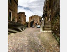 Piazza in acciottolato in un borgo storico, circondata da edifici in pietra e mattoni con due ampie scalinate laterali. Sullo sfondo si vede una casa centrale e alcune auto parcheggiate sotto un cielo nuvoloso.