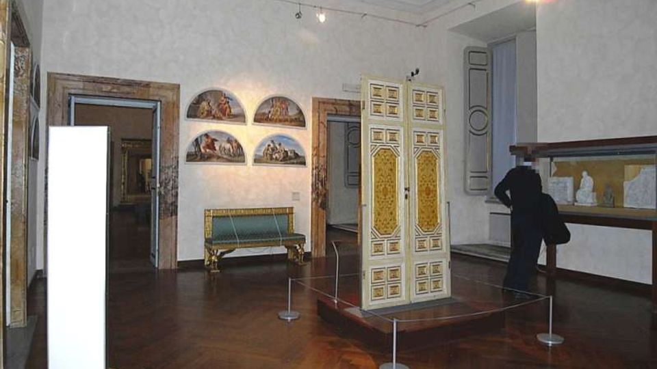 Sala museale con pavimento in parquet e pareti chiare, con un mobile decorato bianco e oro esposto su pedana delimitata da cordoni. Alle pareti si vedono dipinti ad arco, un divanetto e una vetrina con piccole sculture.