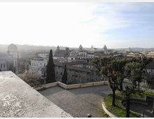 Veduta panoramica di una città storica con tetti in tegole e numerose cupole sullo sfondo, osservata da una terrazza in pietra. In primo piano si vedono una piazzetta lastricata, un muretto perimetrale e alberi lungo il bordo del belvedere.