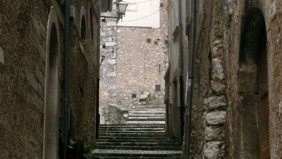 Vicolo stretto in un centro storico, con una lunga scalinata in pietra e pavimentazione a ciottoli tra alti edifici in muratura. In fondo si vede una facciata chiara con finestra rotonda e un lampione a parete lungo il passaggio.