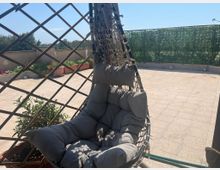 Terrazza pavimentata in piastrelle con una poltrona sospesa intrecciata e imbottita in primo piano. Sullo sfondo ci sono fioriere, una grata in legno e una recinzione coperta di vegetazione lungo il parapetto.