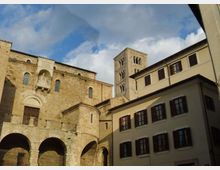 Veduta di una piazza con una grande chiesa romanica in pietra, con portale ad arco, loggia e arcate alla base, affiancata da un campanile quadrato. Sul lato destro si vede un edificio residenziale chiaro a più piani con persiane scure, sotto un cielo parzialmente nuvoloso.