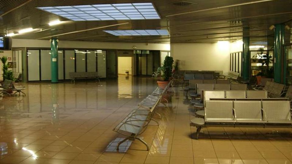 Ampia sala d’attesa interna con pavimento piastrellato lucido, file di sedute metalliche e colonne verdi. Il soffitto è illuminato da luci lineari e pannelli luminosi, con pareti vetrate e alcune piante in vaso lungo i lati.