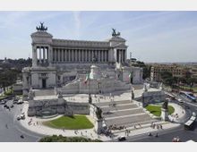 Veduta panoramica del Vittoriano (Altare della Patria) a Roma, grande monumento in marmo bianco con imponente colonnato, scalinate e statue equestri. In primo piano si vede la piazza con aiuole verdi e strade trafficate che circondano il complesso.