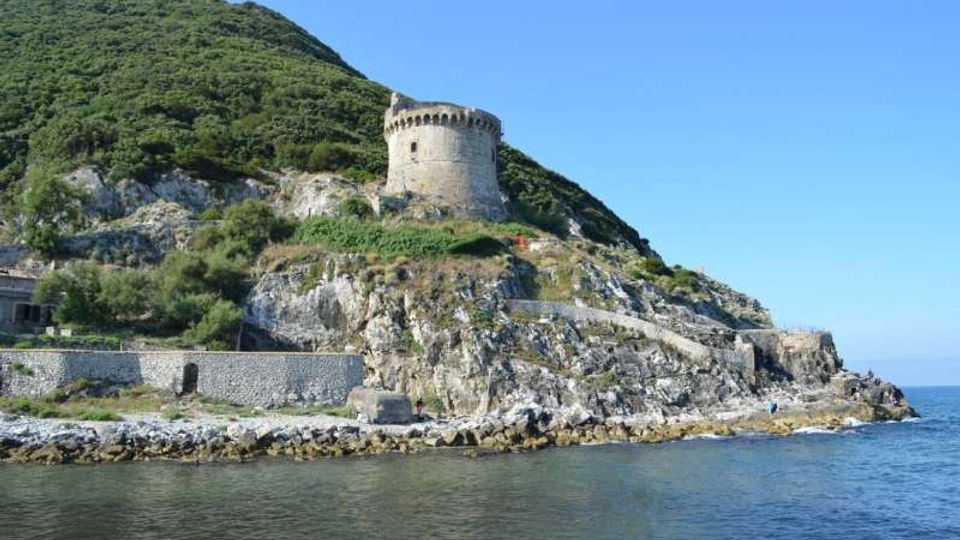 Torre costiera in pietra di forma cilindrica con merlature, situata su una scogliera rocciosa ai piedi di una collina verde. In primo piano si vede il mare con una scogliera e un basso muro di contenimento lungo la riva.