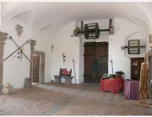 Interno di una sala storica con soffitto a volte bianche e pavimento in mattoni a spina di pesce, con grande portone in legno sul fondo. Alle pareti sono esposte armi e attrezzi antichi, con una piccola panca in pietra e alcuni tavoli coperti da teli.