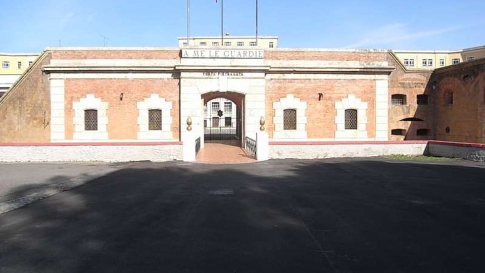 Facciata simmetrica in mattoni con cornici in pietra e ingresso ad arco centrale, sormontato dalla scritta “A ME LE GUARDIE” e da pennoni con bandiera. Davanti si apre un ampio piazzale asfaltato che conduce al varco con cancello verso un cortile interno.