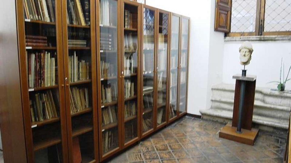 Stanza interna tipo biblioteca o archivio con pavimento in piastrelle e pareti bianche, fiancheggiata da alte librerie in legno con ante in vetro piene di volumi. In fondo si vedono una finestra con inferriate, alcuni gradini in pietra e un busto su piedistallo.