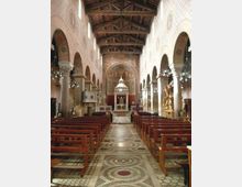 Interno di una chiesa con navata centrale fiancheggiata da arcate e colonne, soffitto a capriate in legno e file di panche in legno. Il pavimento a mosaico con motivi circolari conduce all’altare con ciborio, sullo sfondo di un’abside affrescata.