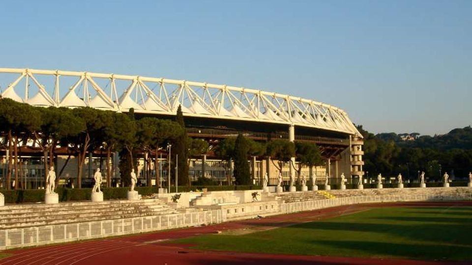 Vista di uno stadio con struttura superiore in traliccio bianco, affiancato da una pista di atletica rossa e un campo erboso. In primo piano si vedono gradinate basse con statue lungo il perimetro e una fila di pini, con colline sullo sfondo.