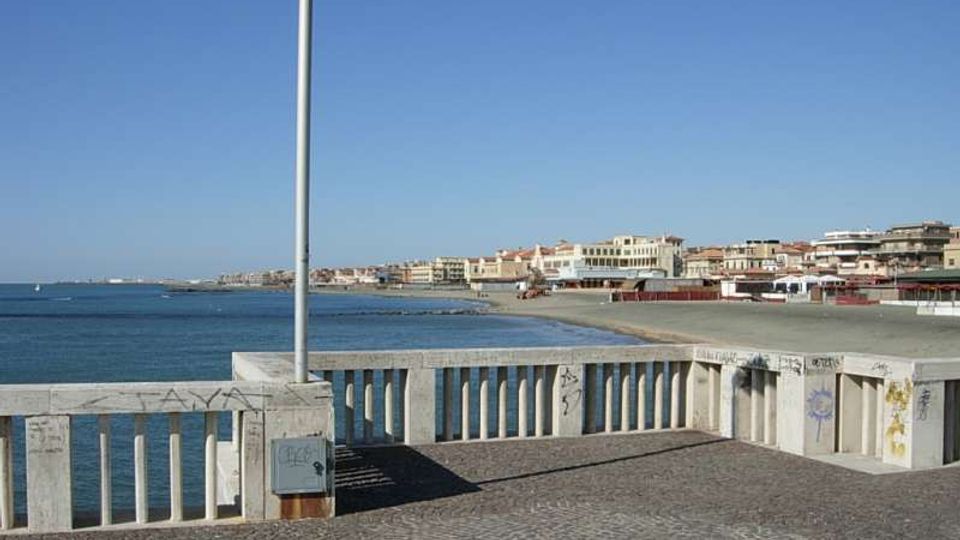 Belvedere sul lungomare con pavimentazione in pietra e balaustra in cemento, con un lampione al centro in primo piano. Sullo sfondo si vede la costa con spiaggia, mare calmo e una fila di edifici lungo la riva sotto un cielo sereno.