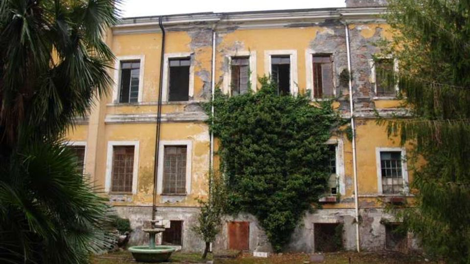 Facciata di un edificio storico a due piani intonacato di giallo, con finestre rettangolari e ampie zone di intonaco scrostato; al centro una grande edera ricopre parte del muro. Davanti si vede un cortile erboso con un piccolo elemento in pietra e vegetazione alta ai lati.