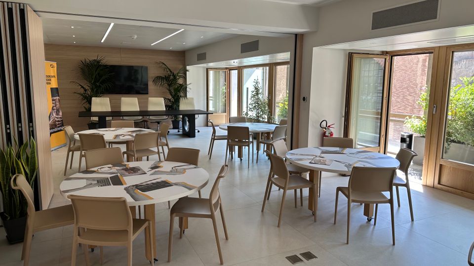 Sala riunioni moderna e luminosa con tavoli rotondi e sedie beige su pavimento chiaro, parete in legno con schermo TV e piante decorative. Grandi porte-finestre con infissi in legno si aprono su una terrazza con verde esterno.