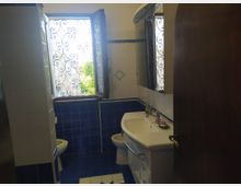 Piccolo bagno con piastrelle blu e bianche, lavabo con mobile e specchio sulla parete. Una finestra aperta con inferriata decorativa lascia entrare luce naturale, con vegetazione visibile all’esterno.