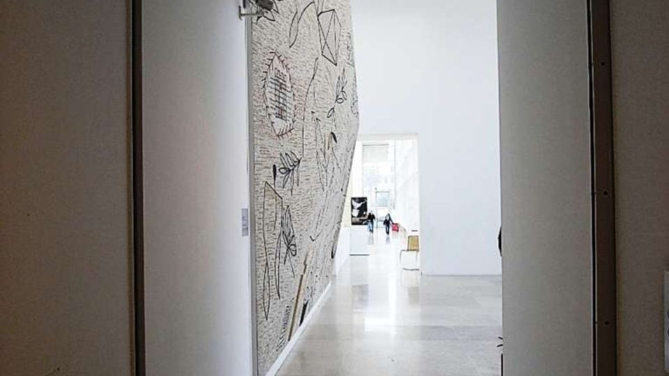 Corridoio interno di un edificio moderno con pareti bianche e pavimento in pietra chiara, illuminato da faretti incassati nel soffitto. Sul lato sinistro si vede una grande parete decorata con disegni lineari, mentre in fondo si apre un ambiente più ampio e luminoso.