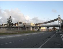 Strada urbana a più corsie con un cavalcavia pedonale moderno dal tetto curvo che attraversa la carreggiata. Sullo sfondo si vedono edifici industriali o di servizio, recinzioni, alberi sparsi e un cielo con nuvole.