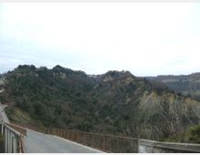 Veduta panoramica di una valle boscosa con colline e pareti di terreno eroso, osservata da un ponte o strada sopraelevata con parapetto metallico in primo piano. Sullo sfondo si vedono crinali e una scarpata chiara sotto un cielo coperto.