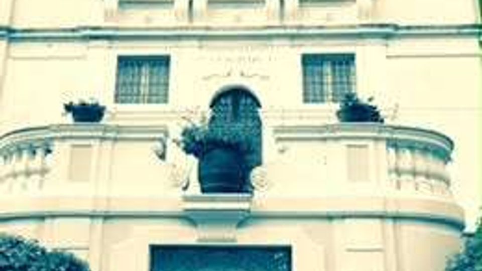 Facciata di un edificio storico a più piani con finestre simmetriche e balconata curvilinea; al centro si vede una nicchia con statua sopra un grande pannello decorativo geometrico, con vegetazione ai lati.