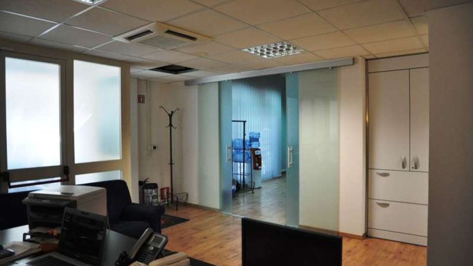 Interno di un ufficio con pavimento in legno, scrivanie con computer e telefono, e grandi finestre con vetri opachi. Sul fondo si vedono una parete vetrata scorrevole che conduce a un corridoio e, sulla destra, armadi bianchi e un appendiabiti.