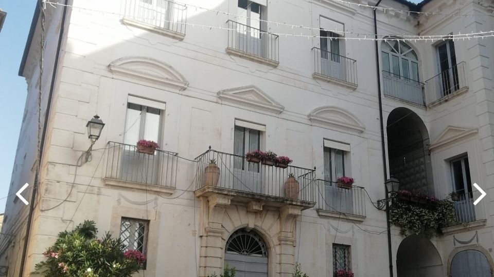 Facciata di un edificio storico intonacato di bianco su una piccola piazza acciottolata, con portale centrale ad arco, balconi con ringhiere e fioriere e finestre simmetriche. A destra si vede un passaggio ad arco che conduce a un cortile interno, con lampioni a muro e fili di illuminazione sospesi sopra.