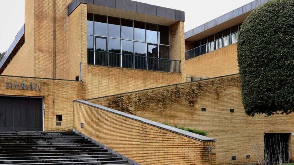 Edificio moderno in mattoni gialli con ampie vetrate e coperture scure, caratterizzato da una grande scalinata e una rampa laterale che conducono all’ingresso. Davanti c’è un’area pavimentata con prato e un grande albero potatu a sfera.