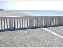 Terrazza sul lungomare con pavimentazione in pietra a ventaglio e balaustra in cemento con pilastrini verticali. Oltre la ringhiera si vede un’ampia spiaggia sabbiosa e il mare calmo fino all’orizzonte.