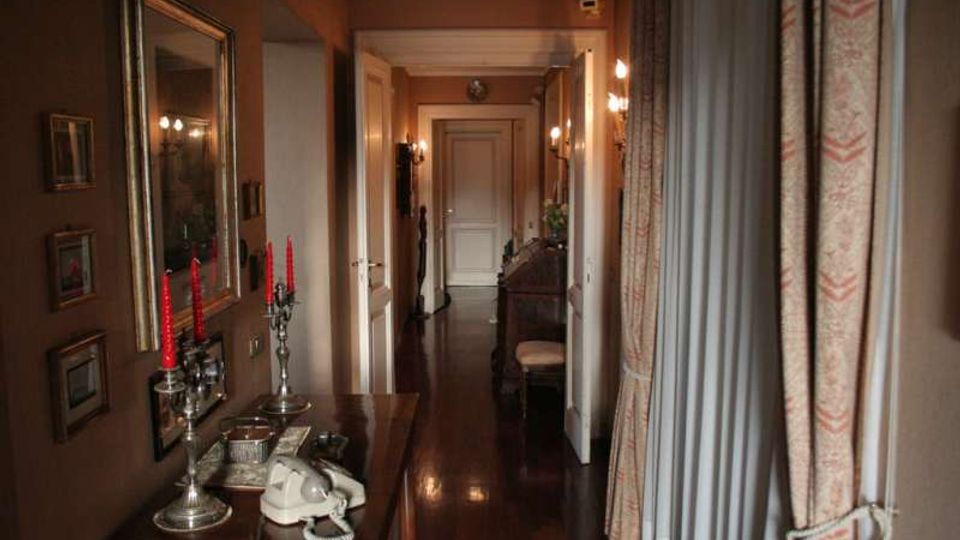 Corridoio interno di una casa con pavimento in legno scuro lucido, pareti color terra e porte bianche lungo il passaggio. A sinistra c’è un mobile con specchio, candelabri e telefono, mentre a destra tende pesanti e lampade a parete illuminano l’ambiente.