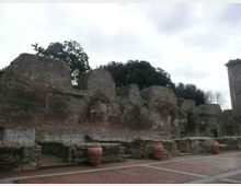 Resti di un antico muro in pietra e mattoni, con ampie porzioni crollate e superfici erose, in un’area archeologica all’aperto. In primo piano si vede una piazza lastricata con muretti bassi e grandi anfore, con alberi sullo sfondo sotto un cielo nuvoloso.