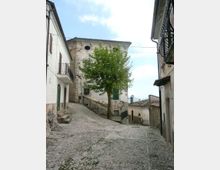Strada in salita lastricata in pietra in un borgo, fiancheggiata da case intonacate con balconi e persiane. Al centro si vede un grande albero davanti a un edificio storico con facciata usurata, con cavi aerei e lampione sullo sfondo.
