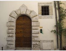 Facciata di un edificio con grande portone ad arco in legno, incorniciato da blocchi di pietra rustica. Accanto al portone c’è una piccola finestra quadrata con inferriata, con tubo di scarico e vegetazione rampicante sul lato destro.