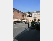 Piazza lastricata in un centro storico, con palazzi color pastello a più piani e un edificio istituzionale sullo sfondo con portale in pietra e piccolo frontone con orologio. In primo piano un lampione in ferro battuto, ai lati tende e bancarelle, ombrelloni e fioriere lungo il bordo della piazza.