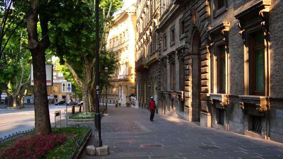 Marciapiede largo in una strada urbana alberata, con aiuole fiorite, lampioni e segnaletica stradale lungo il bordo. Sul lato destro si affacciano edifici storici in muratura con finestre alte e cornici decorative, illuminati dalla luce laterale del sole.