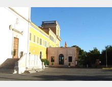 Edificio istituzionale con facciata gialla e bianca, scalinata d’ingresso in pietra e portale laterale, affacciato su un ampio piazzale asfaltato. Al centro si vede un’entrata ad arco con bandiera su un corpo più basso, con alberi e vegetazione sullo sfondo sotto un cielo sereno.