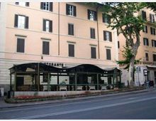 Facciata di un edificio residenziale di più piani color pesca con persiane scure, affacciato su una strada urbana. Al piano terra c’è un ristorante con veranda coperta da una struttura metallica e aiuola lungo il marciapiede, con un grande albero a lato.