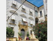 Facciata di un edificio storico intonacato di bianco, con portale ad arco in pietra, balconi con ringhiere e finestre con persiane. Nel cortile antistante si vedono fili di luminarie sospesi, vasi di fiori e alberi ornamentali con fioritura rossa.