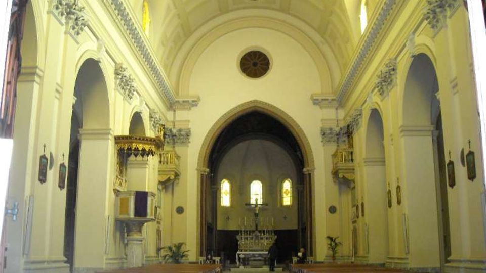 Interno di una chiesa con ampia navata centrale, file di panche in legno e soffitto a volta; ai lati si vedono arcate e decorazioni in stucco. In fondo, un grande arco introduce al presbiterio con altare e finestre ad arco illuminate.