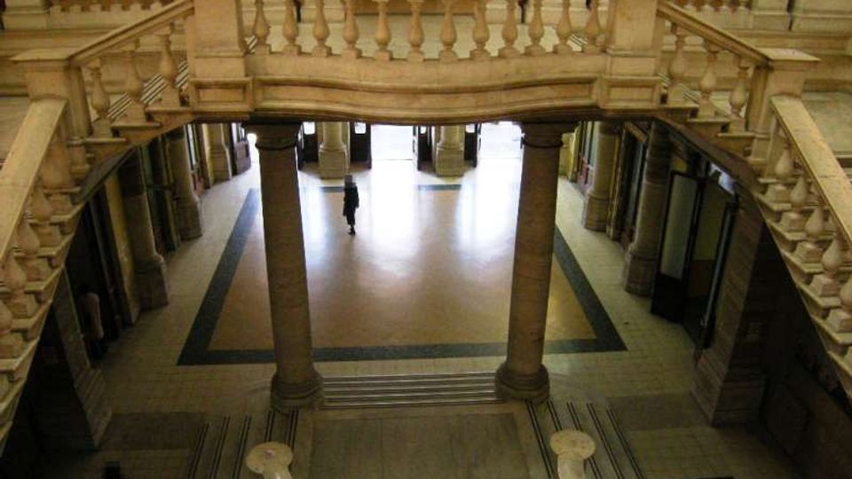 Atrio interno di un edificio storico con ampie scale in pietra e balaustre scolpite che si affacciano su un pianerottolo inferiore con colonne e pavimento a motivi geometrici. Ai lati si vedono corridoi con porte e, sul fondo, un’ampia apertura verso l’esterno.