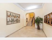 Ingresso condominiale con pavimento in marmo chiaro e pareti color crema, con un grande quadro decorativo sulla sinistra e una fila di cassette postali in legno sulla destra. In fondo, un arco conduce a una scala in pietra con corrimano scuro; accanto è presente una grande pianta d’interno.