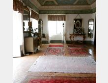Salone interno in stile classico con soffitto decorato a cassettoni, grandi finestre con tende leggere e pavimento in parquet a spina di pesce. La stanza è arredata con consolle intagliate, specchi e tappeti persiani disposti sul pavimento.