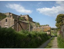 Strada di campagna stretta che costeggia una recinzione metallica, con un complesso di edifici in pietra e tetti in tegole su un pendio erboso. Sullo sfondo cielo azzurro con nuvole e vegetazione rurale ai lati del percorso.