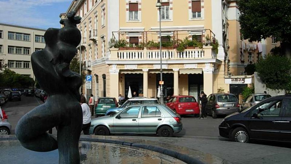 Piazza urbana con una fontana circolare in primo piano e una statua scura al centro, circondata da auto parcheggiate. Sullo sfondo si vede un edificio a più piani color giallo chiaro con balcone e portico ad archi, affiancato da altri palazzi e alberi.