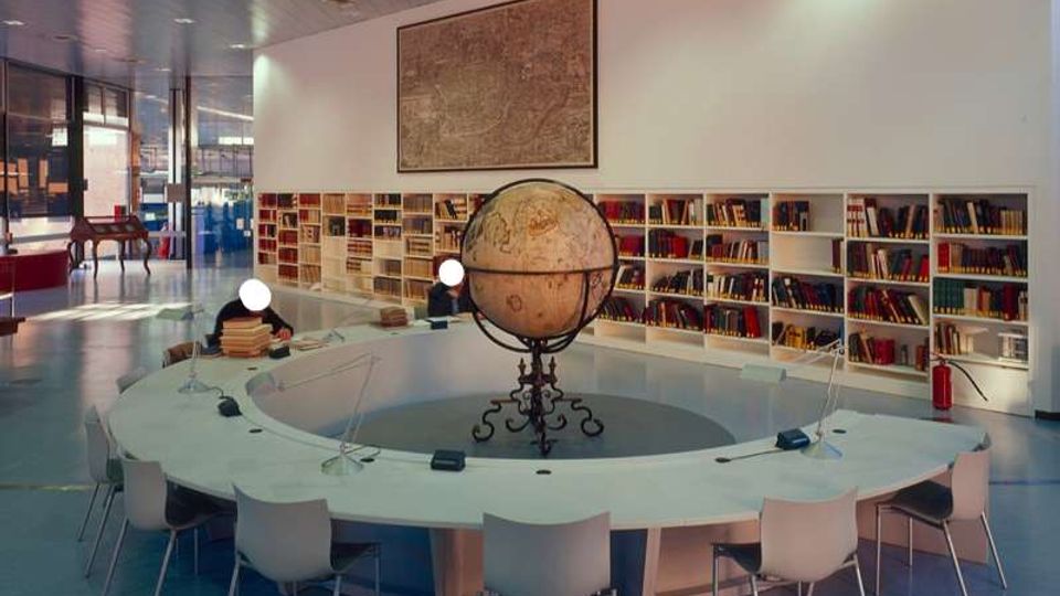 Interno di una biblioteca moderna con grande tavolo circolare bianco e sedie, al centro un mappamondo antico su supporto metallico. Lungo la parete ci sono scaffali bassi pieni di libri e una grande mappa appesa, con ampie vetrate sul lato sinistro.
