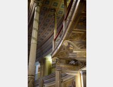 Interno di un edificio storico con colonne alte decorate e un balcone curvo con balaustra in ferro; il soffitto a cassettoni è dipinto con motivi geometrici e dettagli dorati. In primo piano si vedono mensole scolpite con volute e ornamenti che sorreggono la galleria.